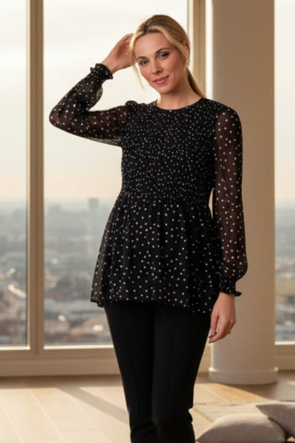 A woman modeling a black long-sleeve sheer mesh top with white polka dots, paired with black leggings, standing in a bright room with large windows overlooking a cityscape.